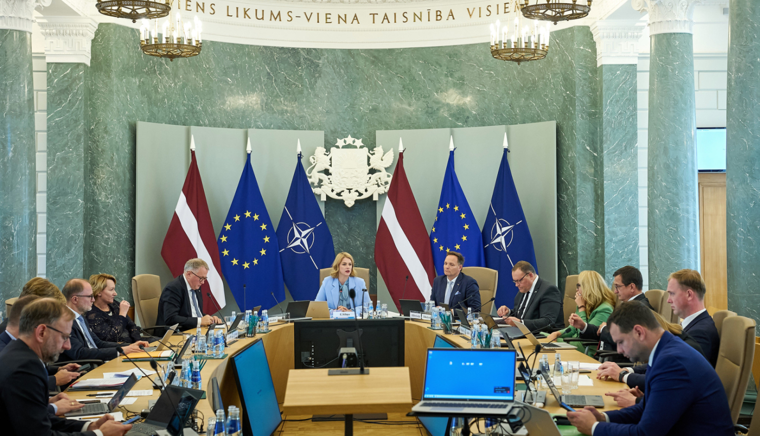 Attēlā redzama Ministru kabineta sēde, kurā visi ministri un Ministru prezidenta sēž pie galda un savstarpēji diskutē, telpas galā pie sienas stāv karogi