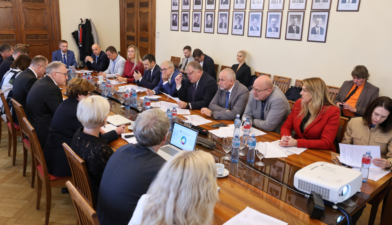 Attēlā garš galds ap kuru no visām pusēm sēž vīrieši un sievietes un savstarpēji sarunājas, uz galda stāv dokumenti, portatīvie datori, ūdens pudeles un glāzes