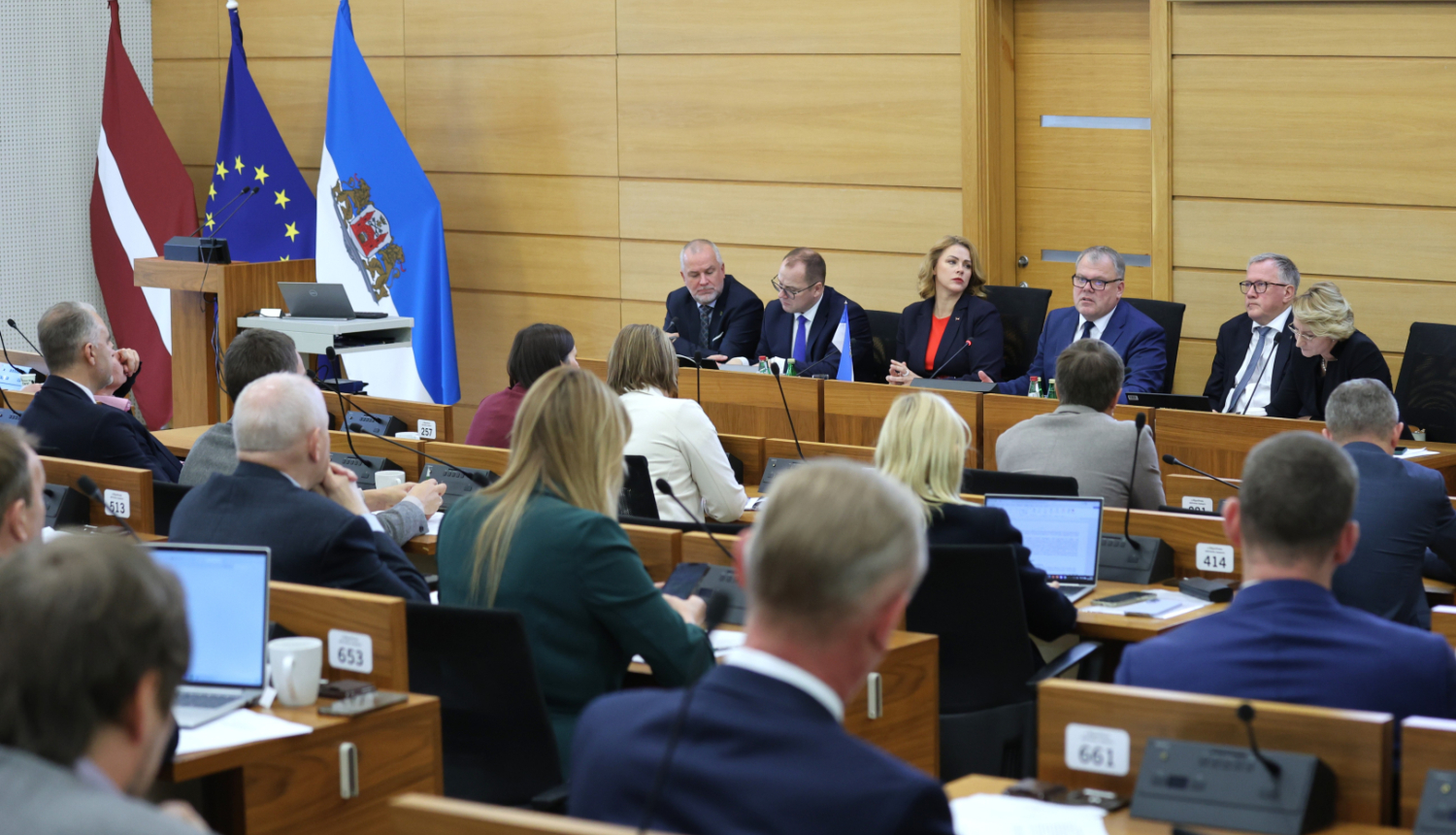 Pie gara galda auditorijas priekšā sēž seši cilvēki, kuri diskutē ar auditoriju, telpas malā stāv Latvijas ES un Rīga domes karogi