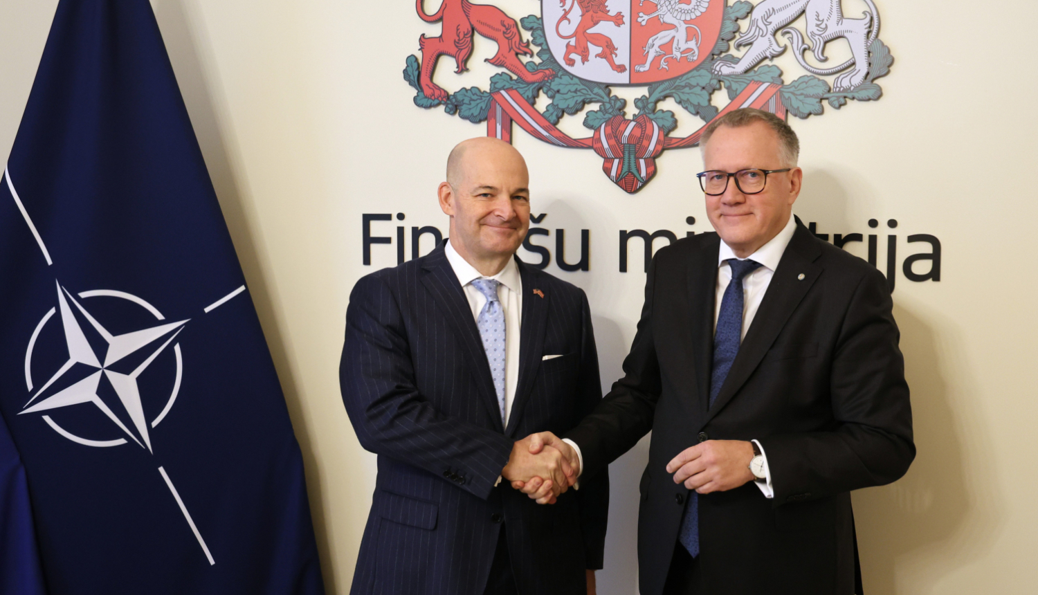 Attēlā uz Finanšu ministrijas ģerboņa fona, sarokojas divi vīrieši uzvalkos, blakus redzams NATO karogs
