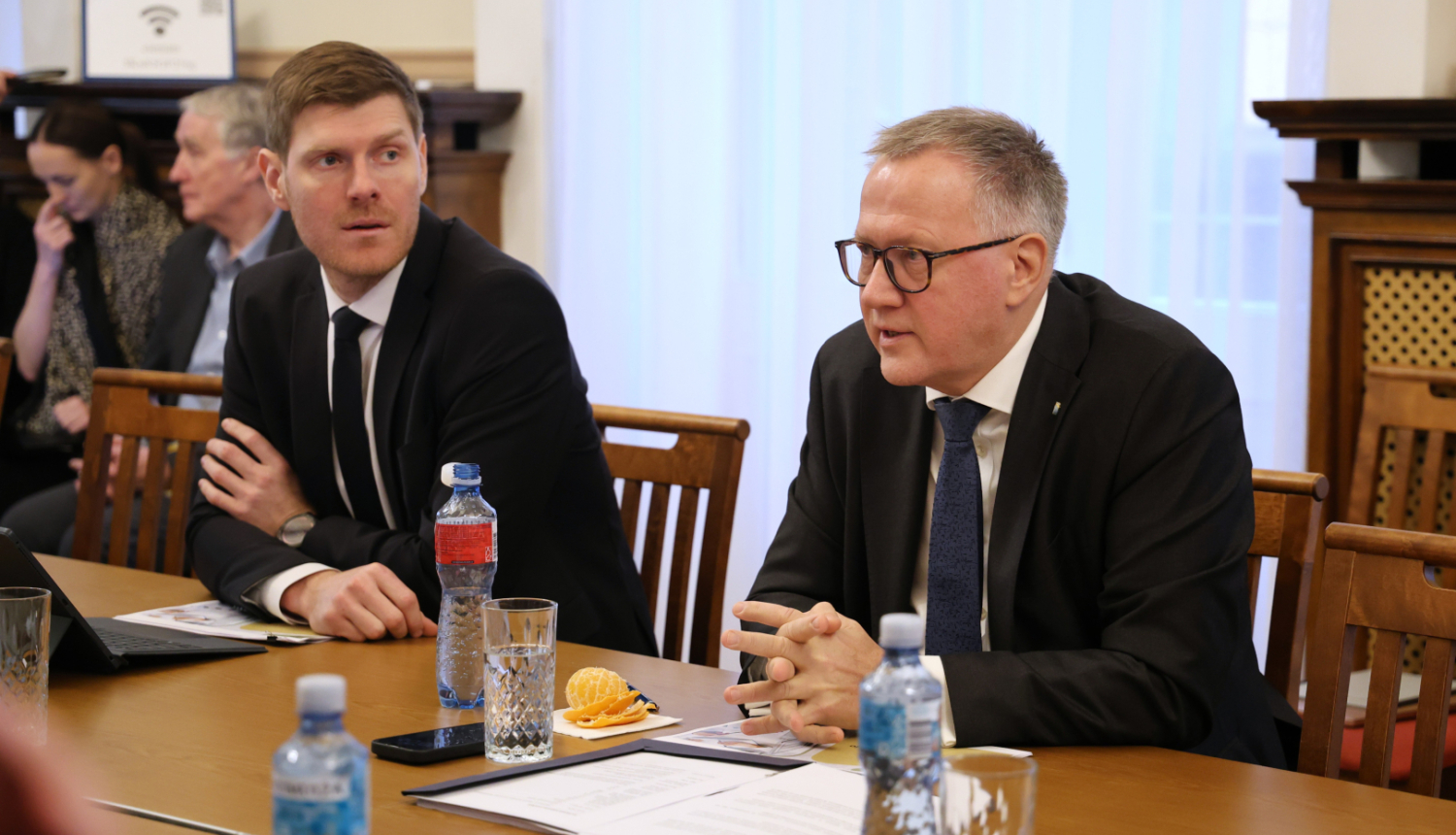 Attēlā pie galda sēžam redzami divi vīrieši uzvalkos, tālumā ir redzami vēl cilvēki sēžam, uz galda stāv dokumenti un ūdens pudeles