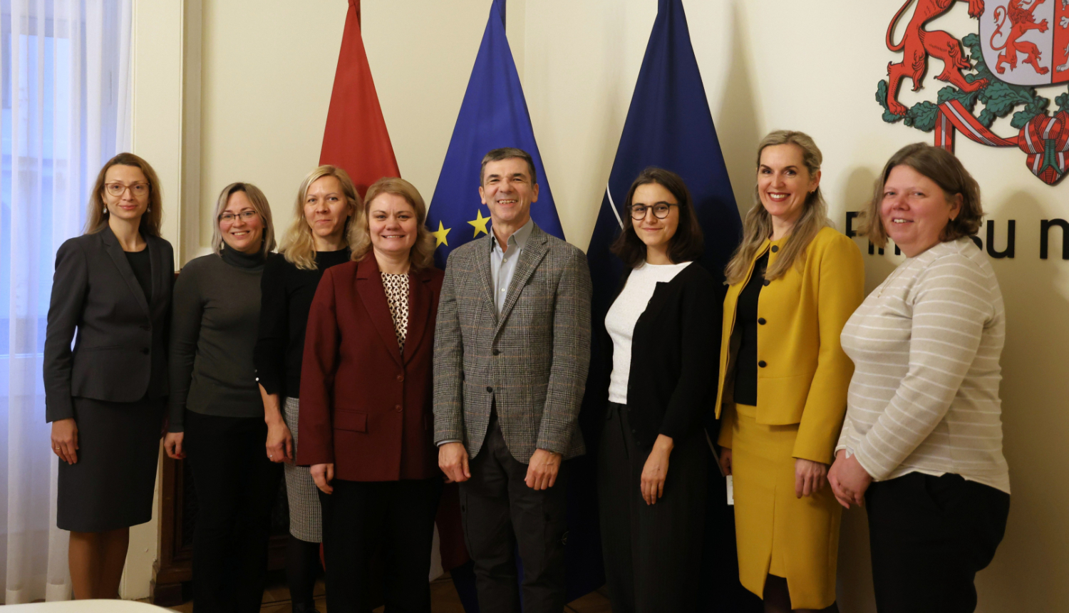 Attēlā uz karogu fona fotografējas rindā stāvošas sievietes un vīrietis, visi smaida