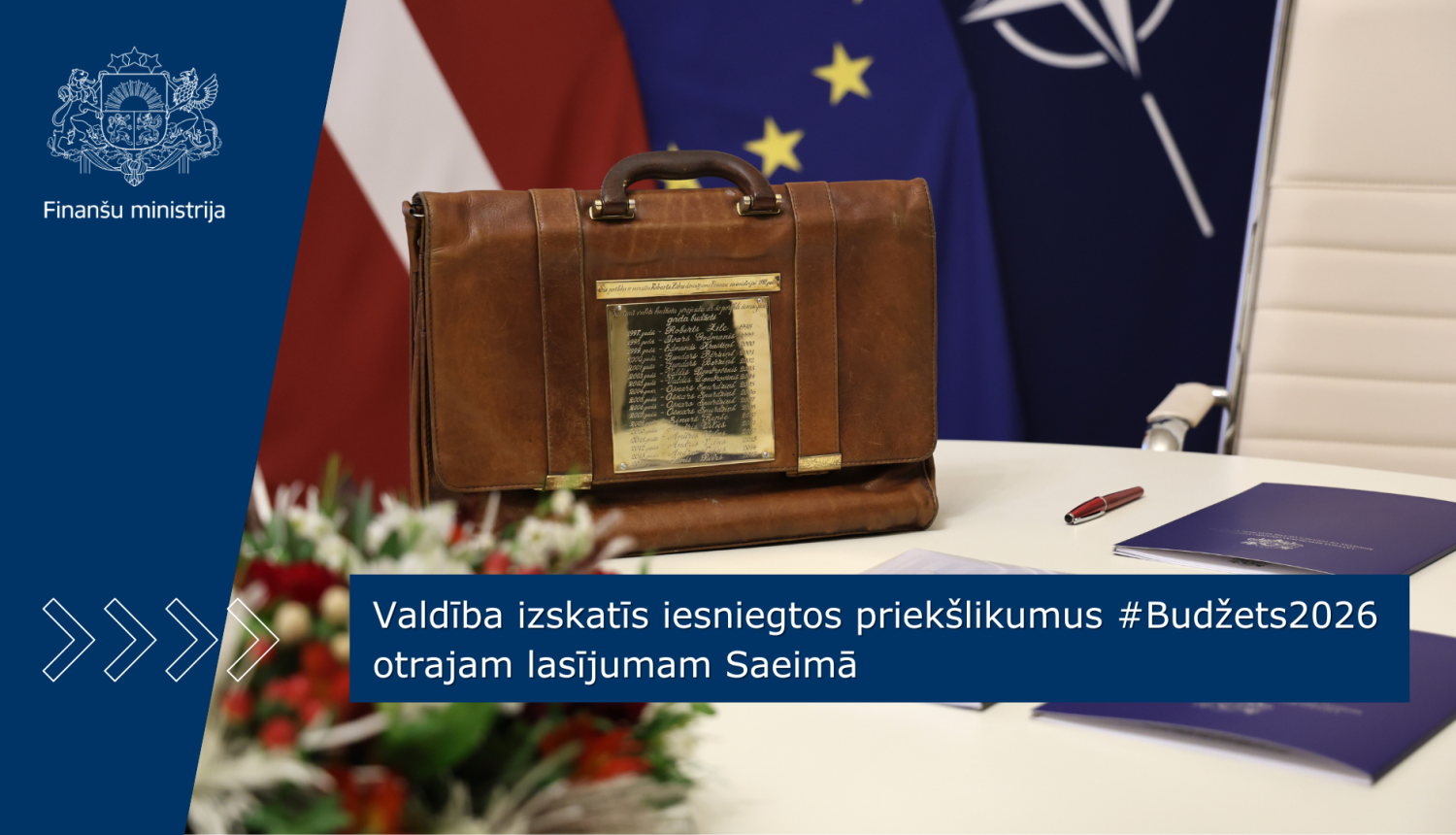 Attēlā redzams tradicionālais budžeta portfelis, kas novietots uz balta galda. Uz galda stāv arī ziedi un zilas dokumentu mapes. Fonā redzami Latvijas, ES un Nato karogi