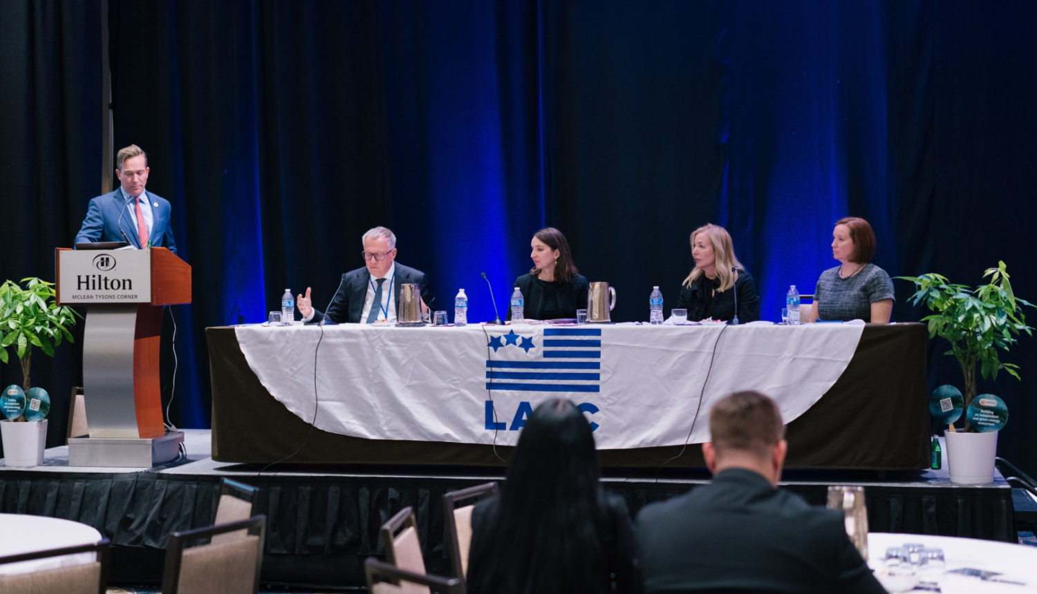 Attēlā pie tribīnes stāv vīrietis zilā uzvalkā, līdzās pie gara galda sēž četri cilvēki - sievietes un vīrieši - runā mikrofonos